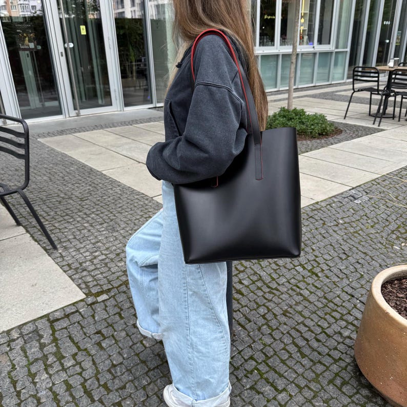 May include: A black tote bag with a smooth, matte finish and long shoulder straps. The straps feature a contrasting red edge detail. The bag has a rectangular, structured shape with clean lines and a minimalist design, suitable for carrying daily items.