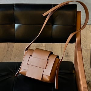 May include: A brown leather crossbody bag with a unique woven design on the front. The bag has a long, adjustable strap and is displayed on a black leather chair. The bag's color is a warm, neutral tone.