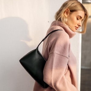 Puede incluir: Un bolso de hombro verde oscuro con forma rectangular y una fina correa negra. El bolso se sostiene contra un su&eacute;ter rosa claro. El fondo es una pared blanca.