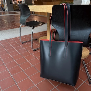 May include: A black tote bag with two long shoulder straps and a vibrant red interior lining. The bag features a smooth, matte exterior finish and thin red piping along the edges of the straps and the top opening, creating a distinct contrast.