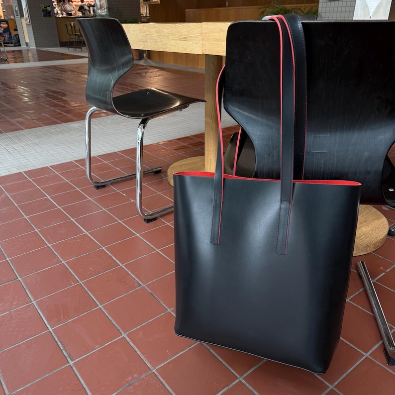 May include: A black tote bag with two long shoulder straps and a vibrant red interior lining. The bag features a smooth, matte exterior finish and thin red piping along the edges of the straps and the top opening, creating a distinct contrast.