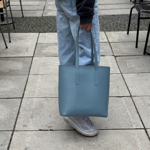May include: A solid, slate blue tote bag with a smooth, matte finish. The bag features a rectangular, structured body with two long, vertical shoulder straps attached to the top edge. The design is simple, clean, and functional in its form.