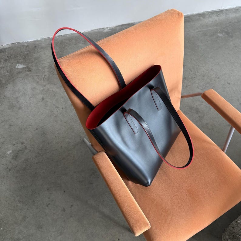 May include: A black tote bag with a smooth exterior and a vibrant red interior lining. The bag features two long, thin shoulder straps that are black on the outside and red on the edges, providing a distinct contrast against the dark main body.