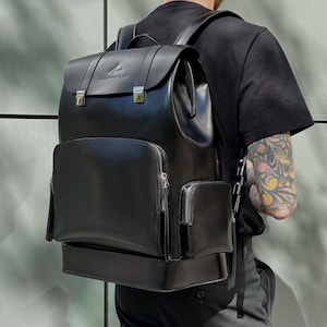 May include: Black leather backpack with multiple zippered compartments and a flap closure. The backpack features silver-colored hardware and the brand name "ALPHON LEW" embossed on the top flap. The bag has adjustable straps and a sleek, modern design.