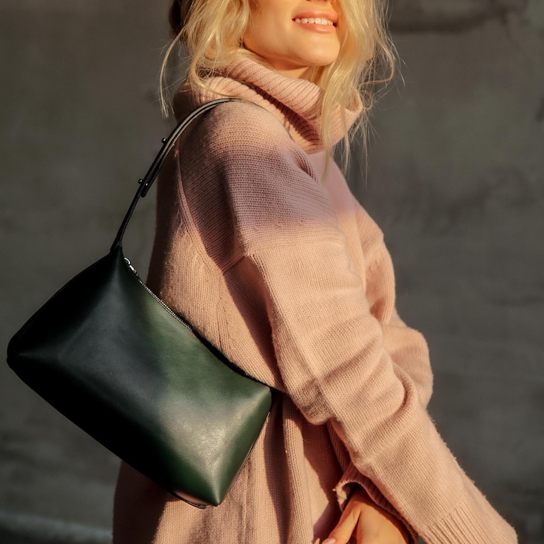Puede incluir: Un bolso de hombro de cuero verde oscuro con cierre de cremallera. El bolso tiene un dise&ntilde;o elegante y minimalista y una correa larga y fina. El bolso se sostiene contra un jersey de cuello alto rosa claro.