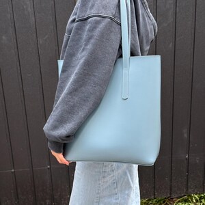 May include: A light blue tote bag with a smooth, matte finish and two vertical shoulder straps. The bag features a rectangular, structured design with clean lines and visible stitching along the strap attachments, providing a simple, functional aesthetic.