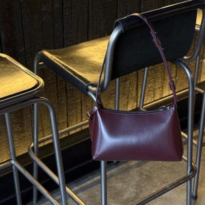 Op de afbeelding: Een bordeauxrode leren schoudertas met een rechthoekige vorm en een lange, dunne riem. De tas hangt aan de achterkant van een zwarte leren stoel met een metalen frame. De achtergrond is een houten muur.