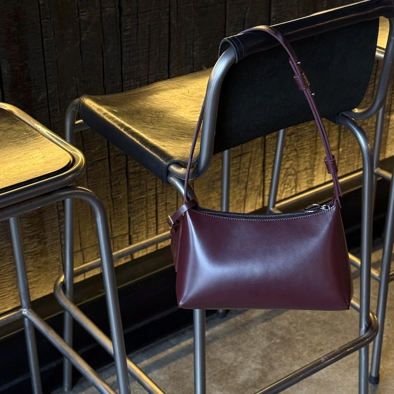 Op de afbeelding: Een bordeauxrode leren schoudertas met een rechthoekige vorm en een lange, dunne riem. De tas hangt aan de achterkant van een zwarte leren stoel met een metalen frame. De achtergrond is een houten muur.