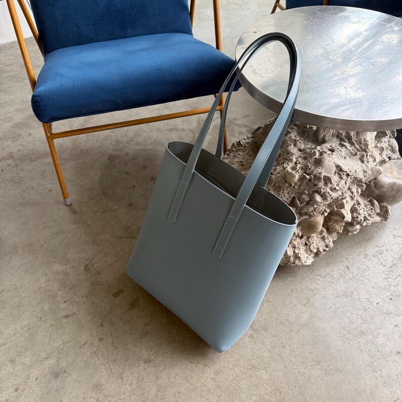 May include: A solid light blue tote bag with a smooth, matte finish. The bag features a tall, rectangular structure with two long, thin shoulder straps attached to the front and back panels, providing a clean and simple design aesthetic.