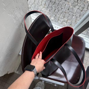 May include: A structured tote bag featuring a smooth black exterior and a vibrant red interior lining. The bag includes two black shoulder straps with red edges and an open top design, revealing a central interior compartment for organization.