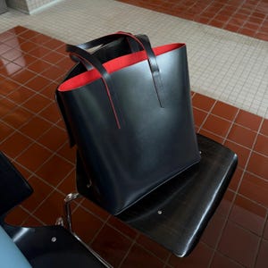 May include: A structured black tote bag with a smooth exterior finish and two vertical handles. The interior lining of the bag is a vibrant red color, which is visible along the top opening and the inner edges of the bag's main compartment.