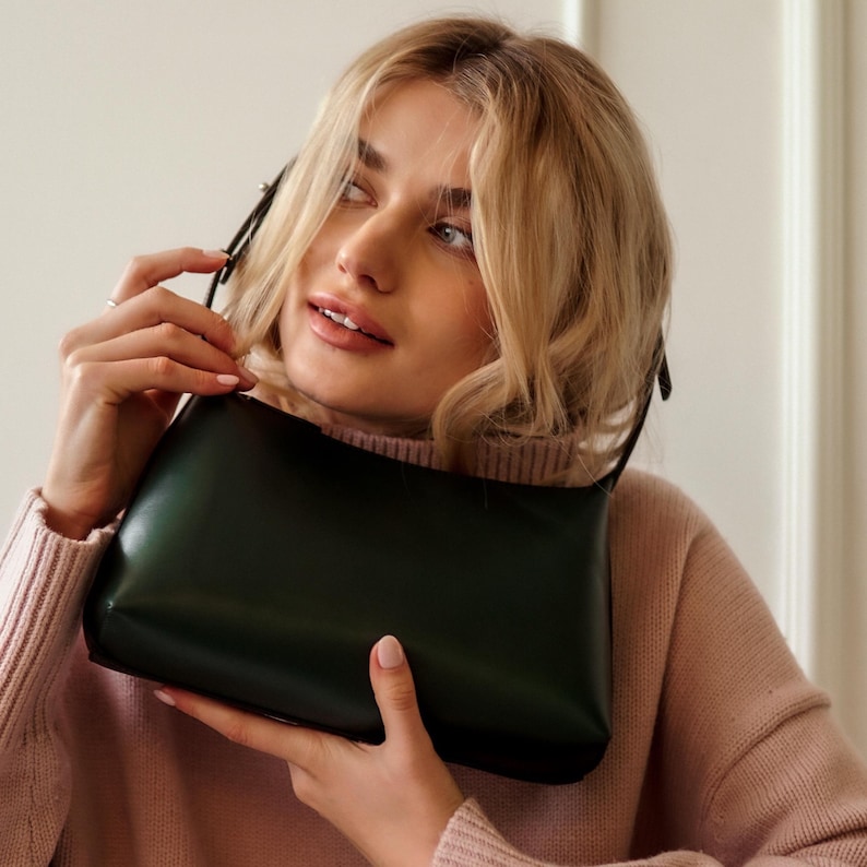Puede incluir: Un bolso rectangular verde oscuro con una correa fina. El bolso lo sostiene una persona que lleva un su&eacute;ter rosa. La superficie lisa del bolso refleja la luz.