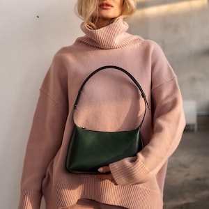 Op de afbeelding: Een vrouw draagt een lichtroze coltrui en bijpassende broek, met een donkergroene leren schoudertas met een zwarte band. De tas heeft een rechthoekige vorm en een ritssluiting.
