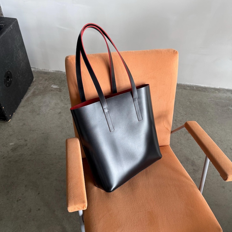 May include: A black tote bag with two long shoulder straps and a contrasting red interior lining. The bag features a smooth, solid-colored exterior surface and a rectangular shape with a wide opening at the top, constructed with clean, straight lines.