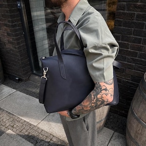 Könnte beinhalten: Eine einfarbige, schwarze, strukturierte Tragetasche mit zwei Tragegriffen und einem abnehmbaren Schultergurt mit silberfarbenen Metallbeschlägen. Die Tasche hat eine glatte, matte Oberfläche und einen rechteckigen Boden.