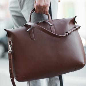 May include: A dark brown leather briefcase featuring dual top handles, a detachable shoulder strap with silver-toned metal hardware, and a zippered main compartment. The front surface displays a subtle embossed logo text reading ALENDE.