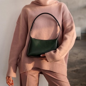 Puede incluir: Un bolso verde oscuro con un asa negra se sostiene contra una persona que lleva un jersey de cuello alto rosa claro y pantalones a juego. El bolso tiene forma rectangular con cierre de cremallera.