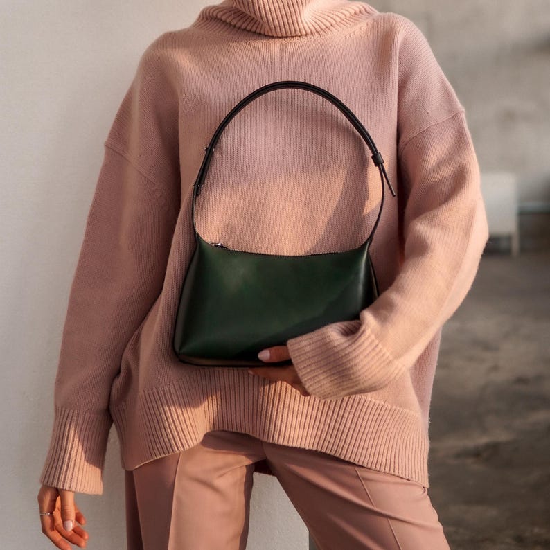 Puede incluir: Un bolso verde oscuro con un asa negra se sostiene contra una persona que lleva un jersey de cuello alto rosa claro y pantalones a juego. El bolso tiene forma rectangular con cierre de cremallera.