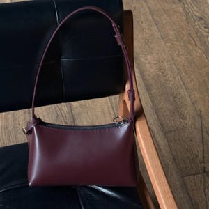 Op de afbeelding: Een bordeauxrode leren schoudertas met een lange, dunne riem. De tas heeft een rechthoekige vorm met een ritssluiting en een kleine, goudkleurige gesp. De tas staat op een zwarte leren stoel en een houten stoel.