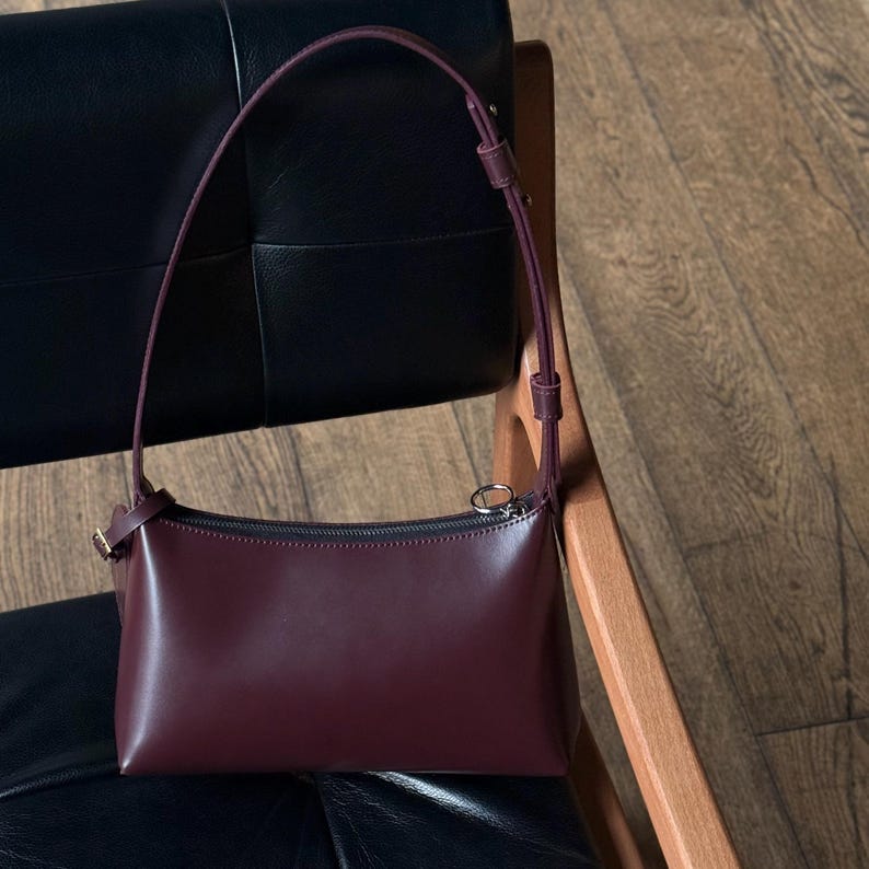 Op de afbeelding: Een bordeauxrode leren schoudertas met een lange, dunne riem. De tas heeft een rechthoekige vorm met een ritssluiting en een kleine, goudkleurige gesp. De tas staat op een zwarte leren stoel en een houten stoel.