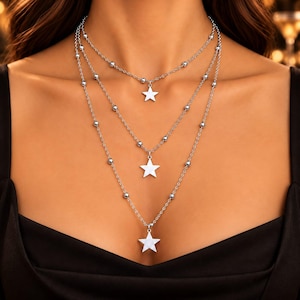May include: A three-layered silver-toned necklace featuring delicate chains adorned with small, evenly spaced metallic beads. Each of the three strands suspends a single, polished five-pointed star charm, creating a cascading effect across the chest.