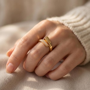 Puede incluir: Un anillo bypass de oro con una banda pulida y lisa que se superpone en el centro. Un extremo de la banda está engastado con una hilera de pequeñas piedras claras de talla redonda, mientras que el otro extremo tiene un acabado dorado sólido.