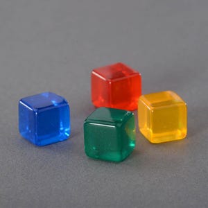 Puede incluir: Cuatro dados translúcidos en azul, rojo, verde y amarillo. Cada dado es un cubo perfecto, con bordes lisos y redondeados. Los dados están dispuestos sobre un fondo gris, siendo cada dado claramente visible.