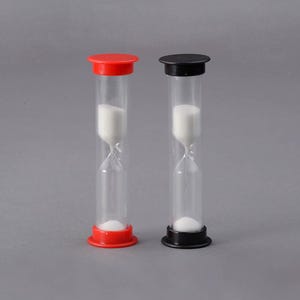 Könnte beinhalten: Zwei Sanduhren mit transparenten Glasröhren, eine mit rotem Deckel und Boden, die andere mit schwarzem. Beide enthalten weißen Sand. Die Sanduhren stehen vor grauem Hintergrund.