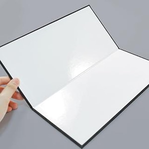 以下が含まれることがあります： 白紙のページが入った黒いノートが開いています。ノートは手で開かれており、光沢のある明るい白い内側が見えます。表紙は黒で、ページはきれいで、書き込みや描画の準備ができています。