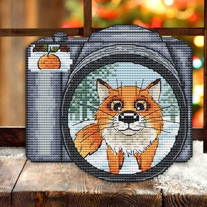 May include: A cross-stitch design of a camera with a fox in the lens. The camera is gray with a small orange fruit on top. The fox is orange and white, set against a snowy background. The image is set against a wooden window.
