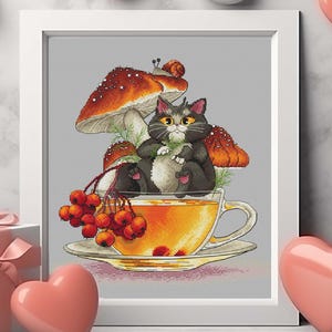 May include: A framed cross-stitch art piece featuring a gray cat in a teacup, surrounded by mushrooms and berries. The artwork is set against a gray background, with decorative pink and white hearts. A gift box with a pink ribbon is also present.