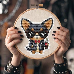 Puede incluir: Bordado de punto de cruz en un aro de madera que presenta un chihuahua de dibujos animados con gafas de sol, pajarita y chaqueta de cuero, sosteniendo una botella. El chihuahua está bordado con hilo marrón, negro y beige. El fondo es neutro.