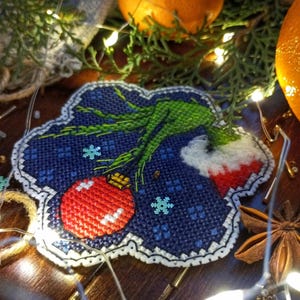May include: A cross-stitched Christmas ornament featuring a green character with a red ornament and snowflakes on a blue background. The ornament has a white scalloped border. Twinkling lights and oranges are in the background.