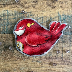 Puede incluir: Un parche hecho a mano con forma de pájaro rojo en punto de cruz, con detalles blancos y amarillos. El pájaro tiene una máscara blanca y un rayo amarillo. El parche está sobre una superficie de madera con cuentas dispersas.