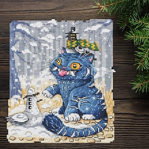 May include: Cross-stitch art featuring a blue cat with yellow eyes, a snowman, and a bird in a winter scene. The cat wears a hat and scarf. Snowflakes fall in the background. The artwork is on a square, textured canvas.