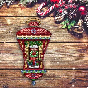 May include: A red and green cross-stitch Christmas lantern with a gold top and a blue and gold bottom. The lantern features a window with a wreath and presents. The background is a wooden surface with Christmas decorations.