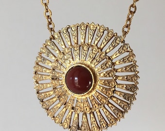 Puede incluir: Un collar de oro con un gran colgante redondo que presenta un diseño detallado de rayos de sol con una piedra preciosa roja en el centro.