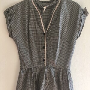 Op de afbeelding: Een vintage zwart-witte geruite jumpsuit met korte mouwen en een V-hals. De jumpsuit heeft een knoopsluiting aan de voorkant, een getailleerde taille en delicate witte kanten afwerking langs de halslijn. De mouwen zijn voorzien van kleine knopen.