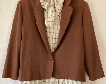 May include: A brown blazer with a single button closure and a white blouse with a checkered pattern underneath. The blouse has a bow tie at the neck.