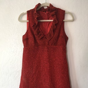 May include: A sleeveless, red dress with a textured surface and a ruffled neckline. The dress has a V-neck and a fitted bodice that flares out slightly at the waist. The dress is hanging on a beige hanger against a white wall.