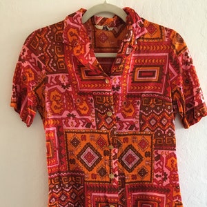 Puede incluir: Una camisa de manga corta con botones y un patrón geométrico en tonos naranja, rosa y marrón. La camisa tiene cuello y cierre de botones.