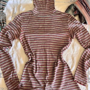 May include: A striped turtleneck sweater in shades of red, white, and green. The sweater is made of a soft, stretchy fabric and has a fitted silhouette.
