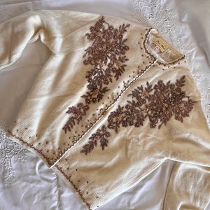 May include: A cream-coloured cardigan with detailed brown floral embroidery and bead embellishments. The sweater has long sleeves and a button-up front. The embroidery is concentrated on the front and sleeves, creating a decorative design.