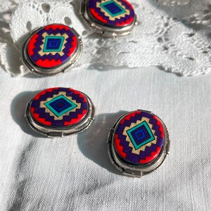 May include: Four oval-shaped earrings with a silver setting and a colourful geometric design. The design features red, purple, blue, green, and gold colours.