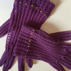 May include: A pair of purple lace gloves with gold studs. The gloves are folded in half, with the fingertips of one glove tucked into the other.