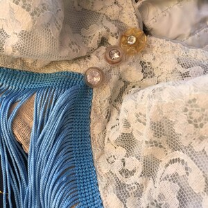 Puede incluir: Primer plano de un tejido de encaje blanco con ribete de flecos azules y beige. Hay tres botones decorativos, uno dorado y dos transparentes, en la tela.