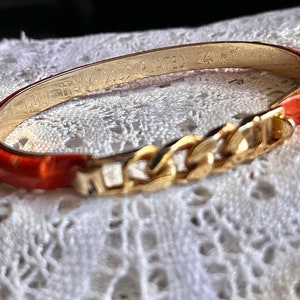 May include: A gold-plated bangle bracelet with a red and orange resin inlay. The bracelet is engraved with "Made in Italy Gold Plated 14 KT".