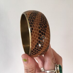 Könnte beinhalten: Ein braunes und schwarzes gemustertes Armreif-Armband mit einem goldenen Metallband.