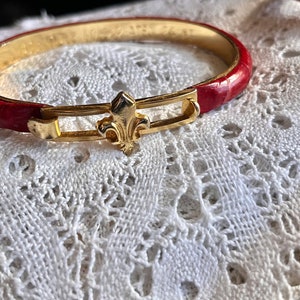 May include: A red leather bangle bracelet with a gold fleur-de-lis clasp.