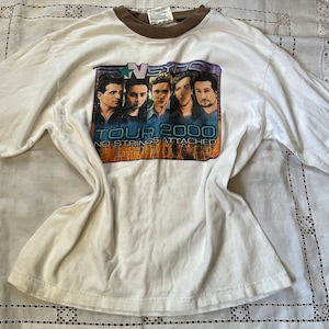 Op de afbeelding: Wit T-shirt met een bruine kraag en een afbeelding van de band *NSYNC op de voorkant. De afbeelding zegt "Tour 2000 No Strings Attached".